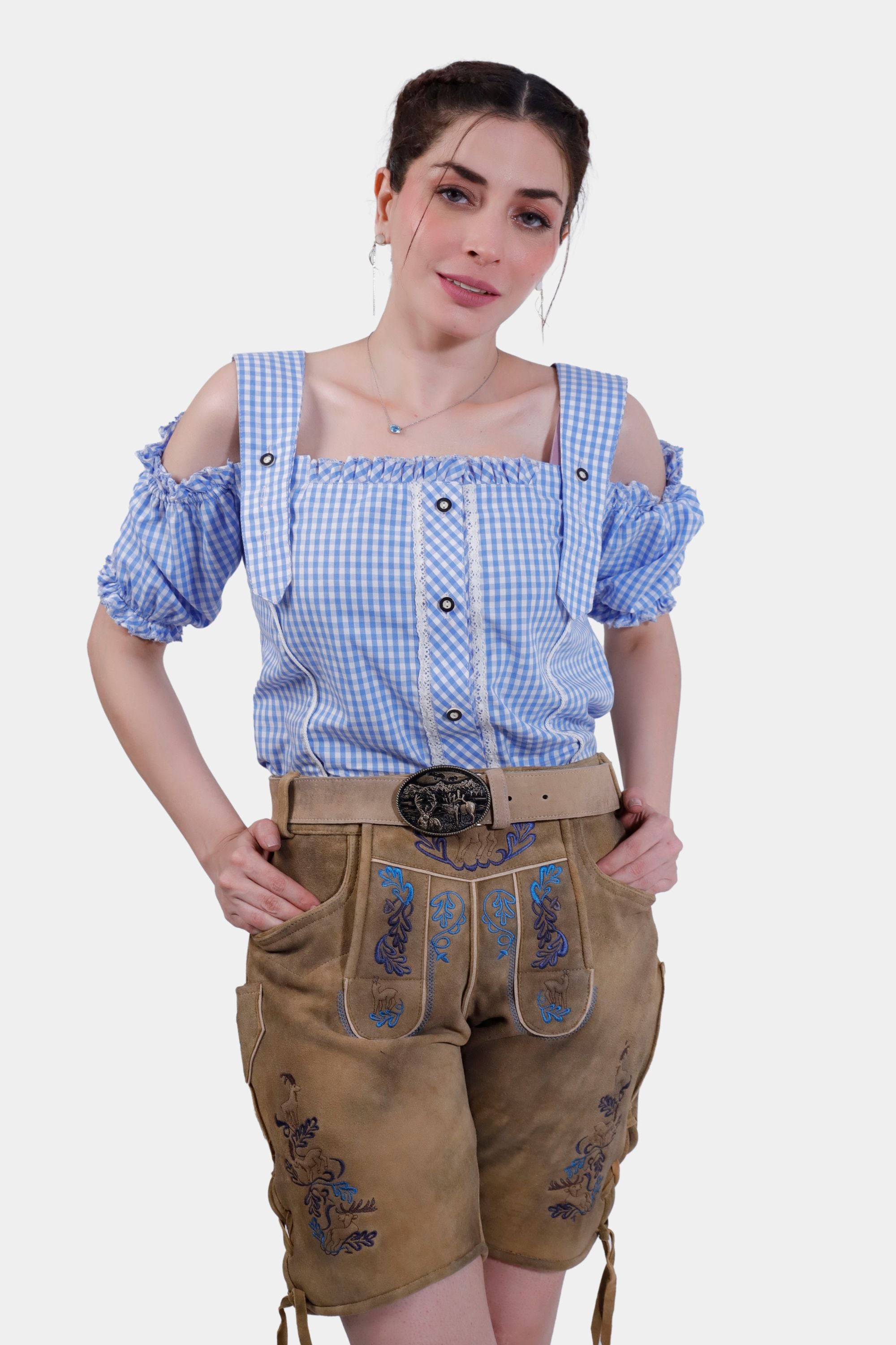 A woman wearing Clara Schumann Lederhosen in Bluebell Caramel, paired with a blue and white checkered blouse. The Lederhosen feature intricate blue embroidery on the bib and legs.