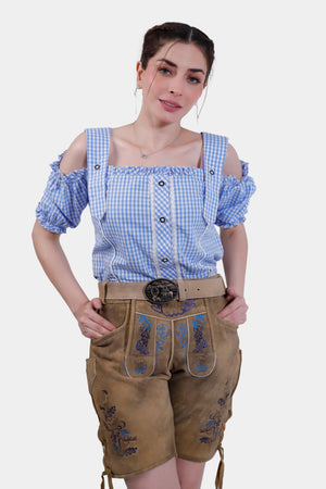 A woman wearing Clara Schumann Lederhosen in Bluebell Caramel, paired with a blue and white checkered blouse. The Lederhosen feature intricate blue embroidery on the bib and legs.