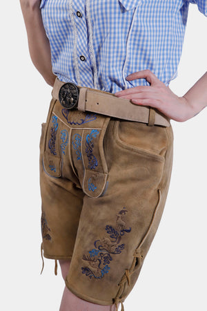 A woman wearing tan leather lederhosen with intricate blue embroidery, featuring side pockets and lace-up details at the hem, standing with one hand on her hip.