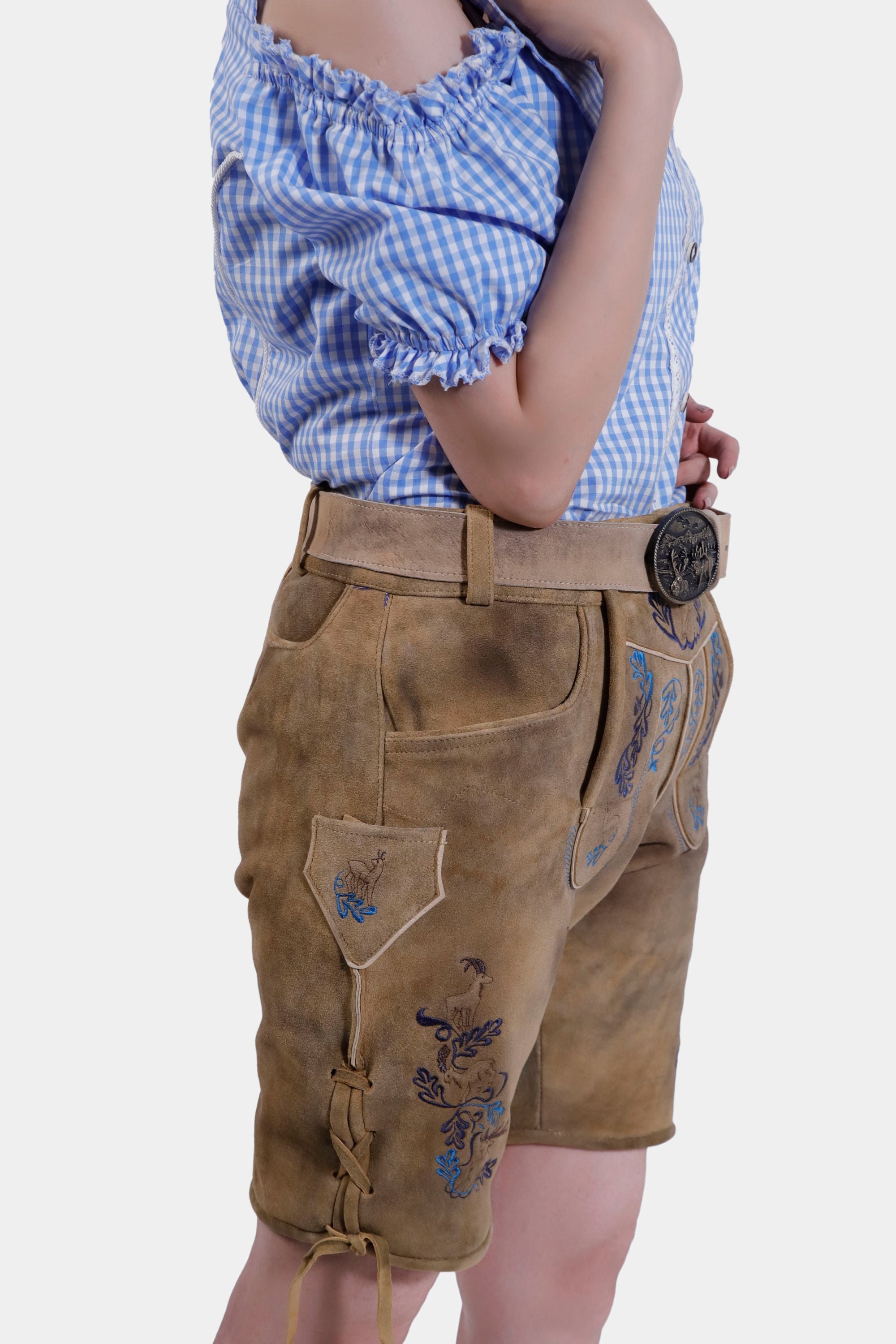 Side view of the Clara Schumann Lederhosen showing the side knife pocket and intricate embroidery on the legs. The model pairs it with a blue and white checkered blouse.