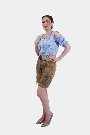 A full-body shot of a woman wearing a blue and white checkered blouse and tan leather lederhosen with blue embroidery, standing confidently with her hands on her hips.