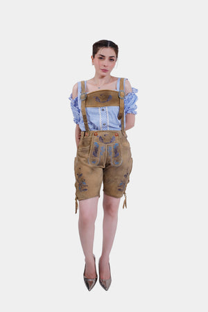 A woman wearing a blue and white checkered blouse and tan leather lederhosen with blue embroidery and detailed buckles, standing with her hands behind her back.