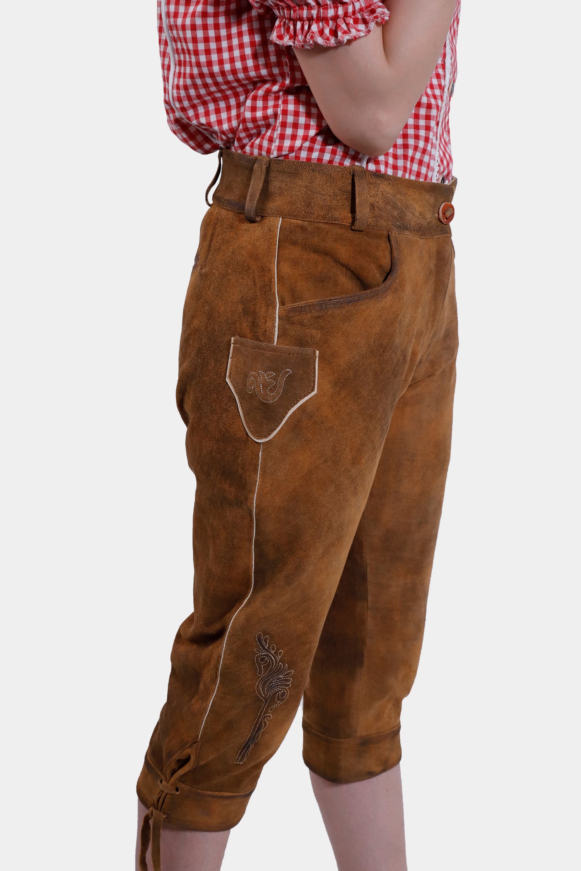 A side view of the brown leather lederhosen, focusing on the embroidery and pocket details. The woman is wearing a red checkered blouse, with her hand resting on her hip.