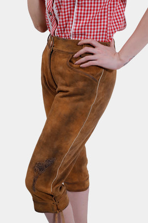 Close-up side view of a woman wearing light brown knee-length Lederhosen with simple embroidery on the leg. The woman is dressed in a red checkered blouse, her hand resting on her hip.