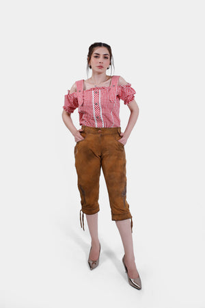 A full-body view of a woman wearing a red checkered off-shoulder blouse and brown leather lederhosen. She stands confidently with her hands in her pockets, wearing metallic high heels.