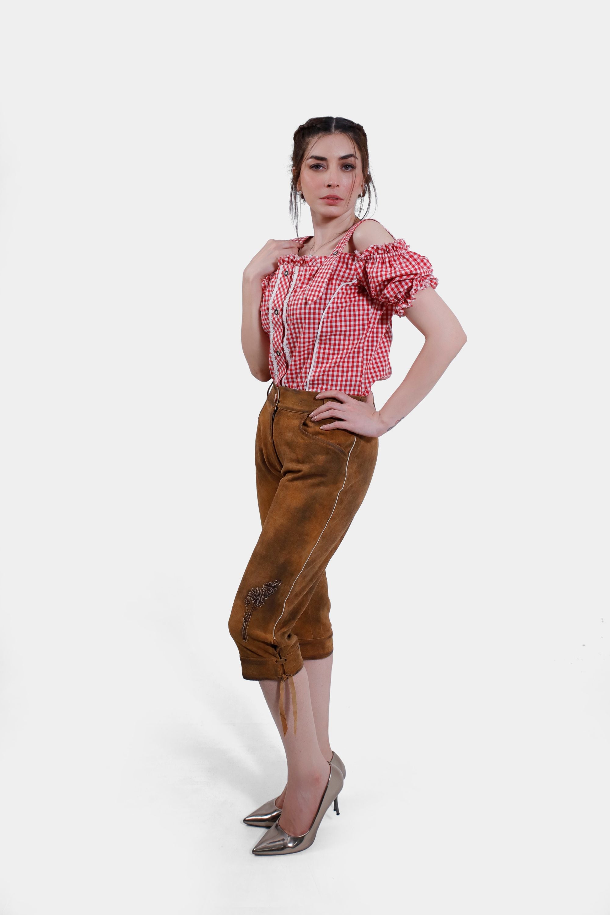Full-body image of a woman wearing light brown knee-length Lederhosen with minimal embroidery, paired with a red checkered blouse. She stands confidently with one hand resting on her hip.