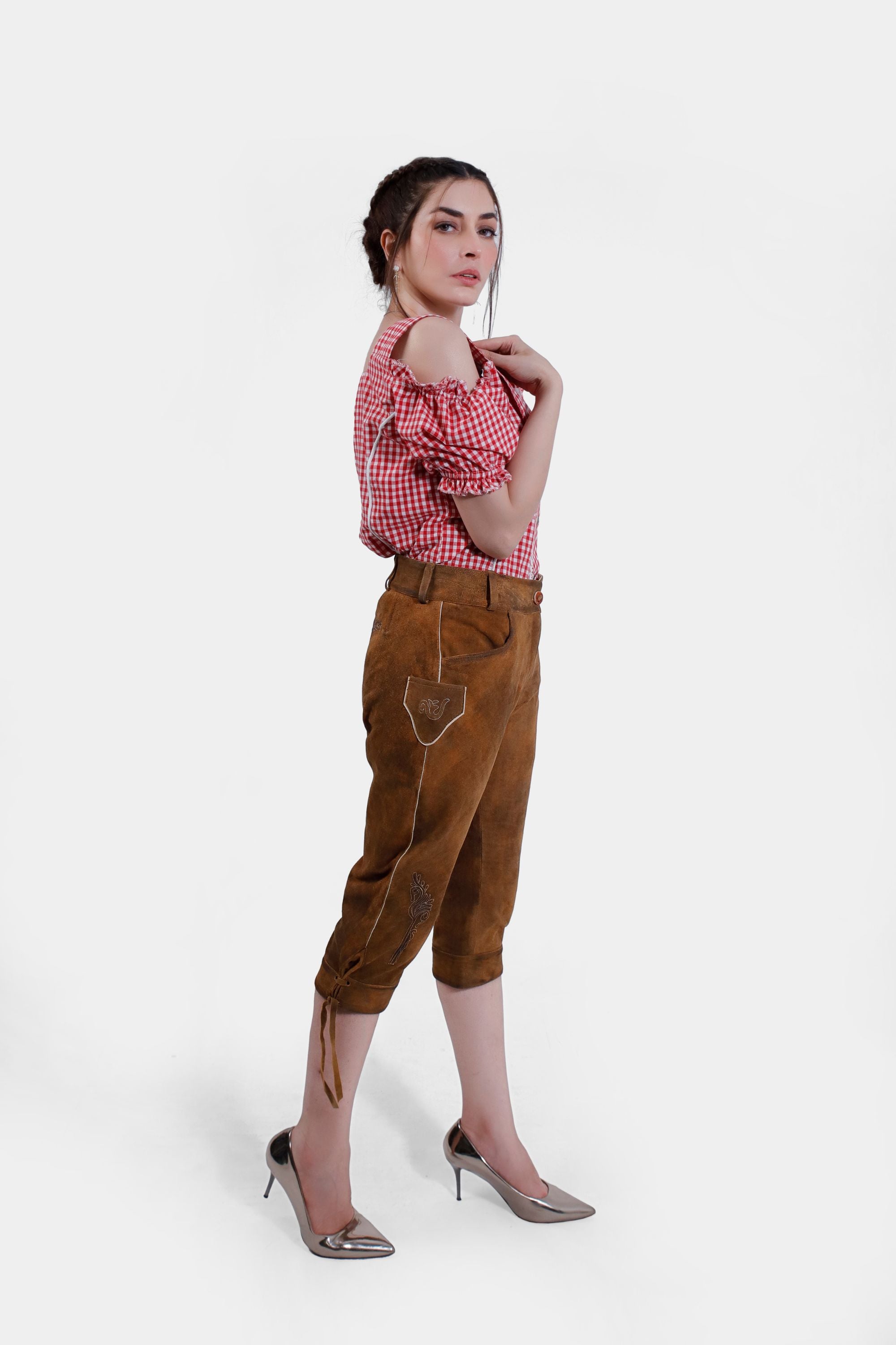 Side profile of a woman wearing light brown knee-length Lederhosen with delicate embroidery and a red checkered blouse. She stands in a slightly turned pose, with one hand resting on her shoulder.