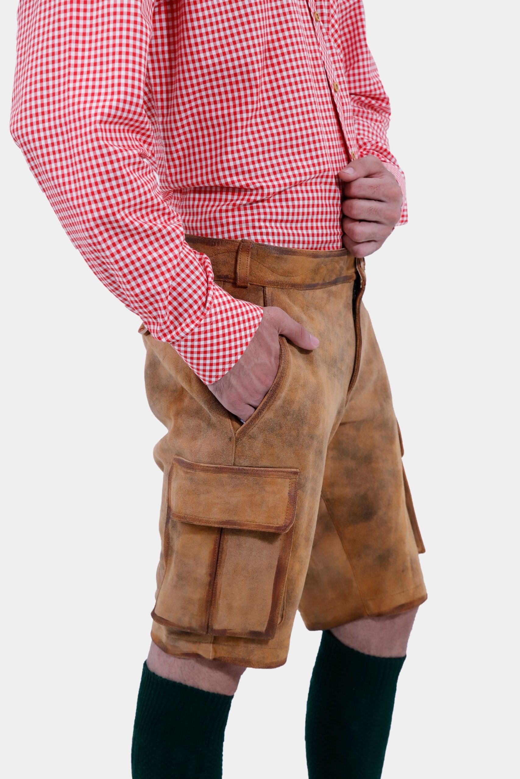 closed view of a man with a beard is standing in a three-quarter view against a white background, wearing a red and white checkered shirt and brown leather lederhosen. He has his right hand in the pocket of the lederhosen, and is also wearing dark green knee-high socks.
