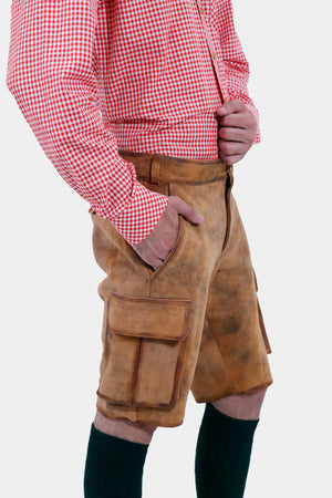closed view of a man with a beard is standing in a three-quarter view against a white background, wearing a red and white checkered shirt and brown leather lederhosen. He has his right hand in the pocket of the lederhosen, and is also wearing dark green knee-high socks.