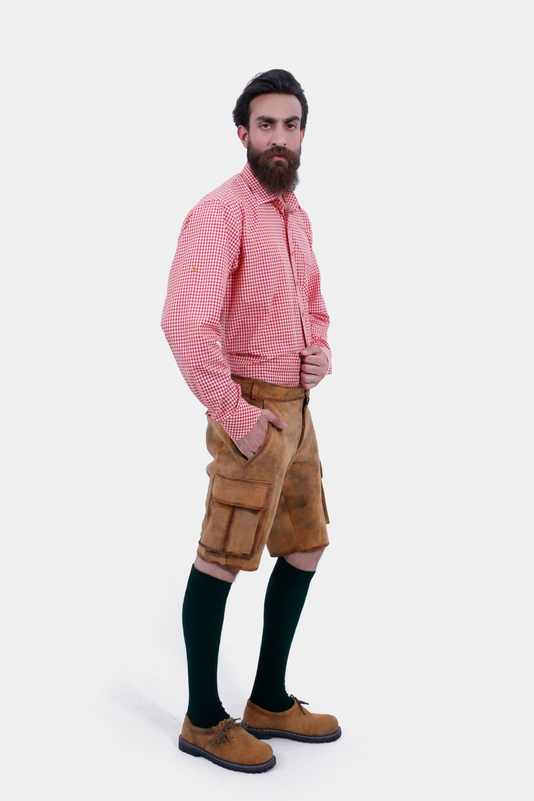 A man with a beard is standing in a three-quarter view against a white background, wearing a red and white checkered shirt and brown leather lederhosen. He has his right hand in the pocket of the lederhosen, and is also wearing dark green knee-high socks and brown shoes.