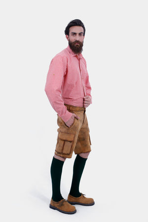 A man with a beard is standing in a three-quarter view against a white background, wearing a red and white checkered shirt and brown leather lederhosen. He has his right hand in the pocket of the lederhosen, and is also wearing dark green knee-high socks and brown shoes.