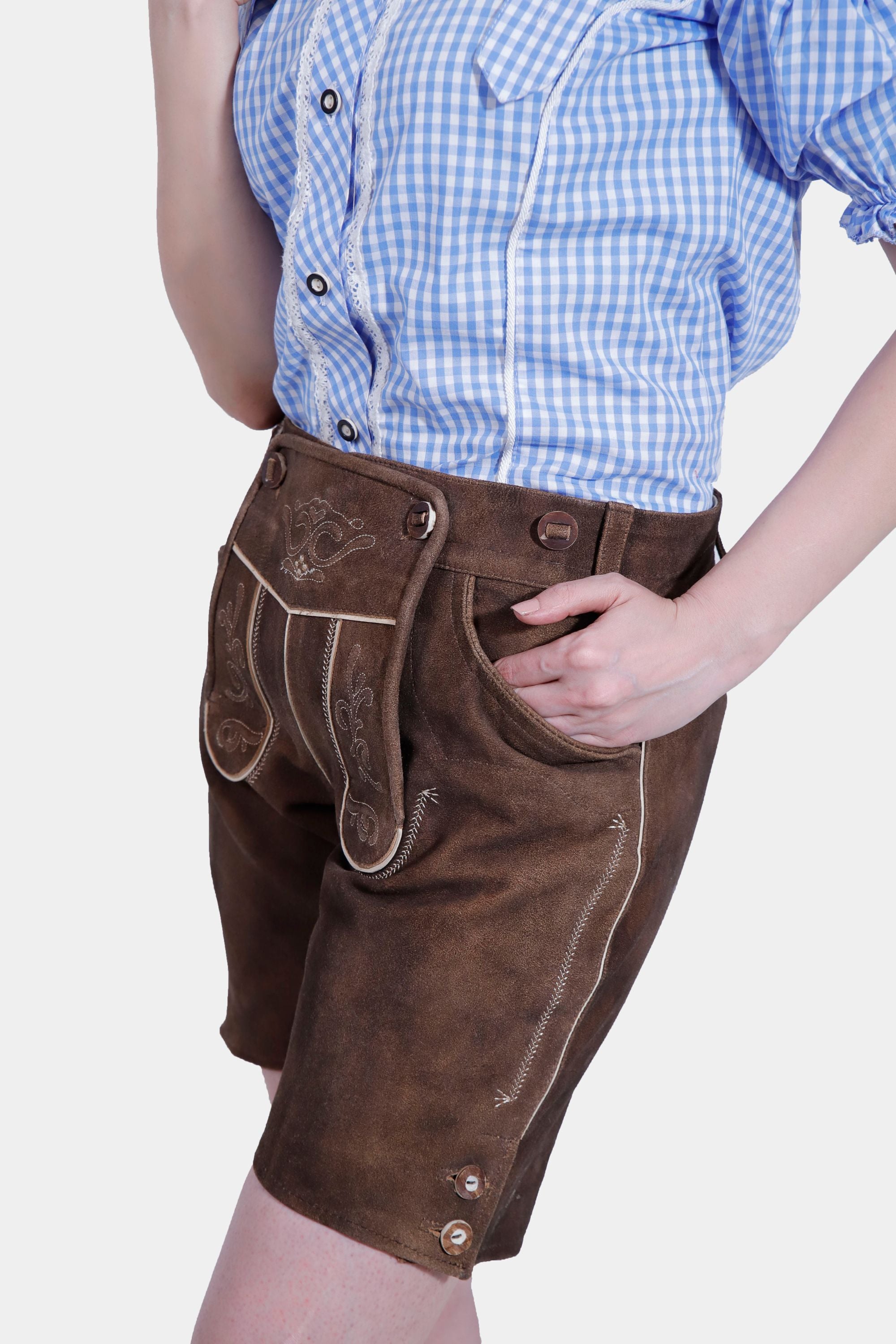 Close-up of a model's outfit highlighting the detailed embroidery on brown leather lederhosen shorts paired with a blue checkered blouse. The model's hand is casually placed in the pocket.