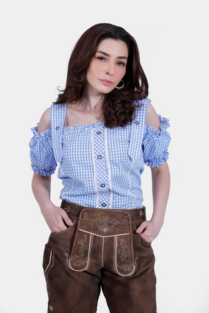 Close-up of a woman in a blue and white checkered off-shoulder blouse and brown short lederhosen. She has her hands in her pockets, highlighting the detailed embroidery and design of the lederhosen.