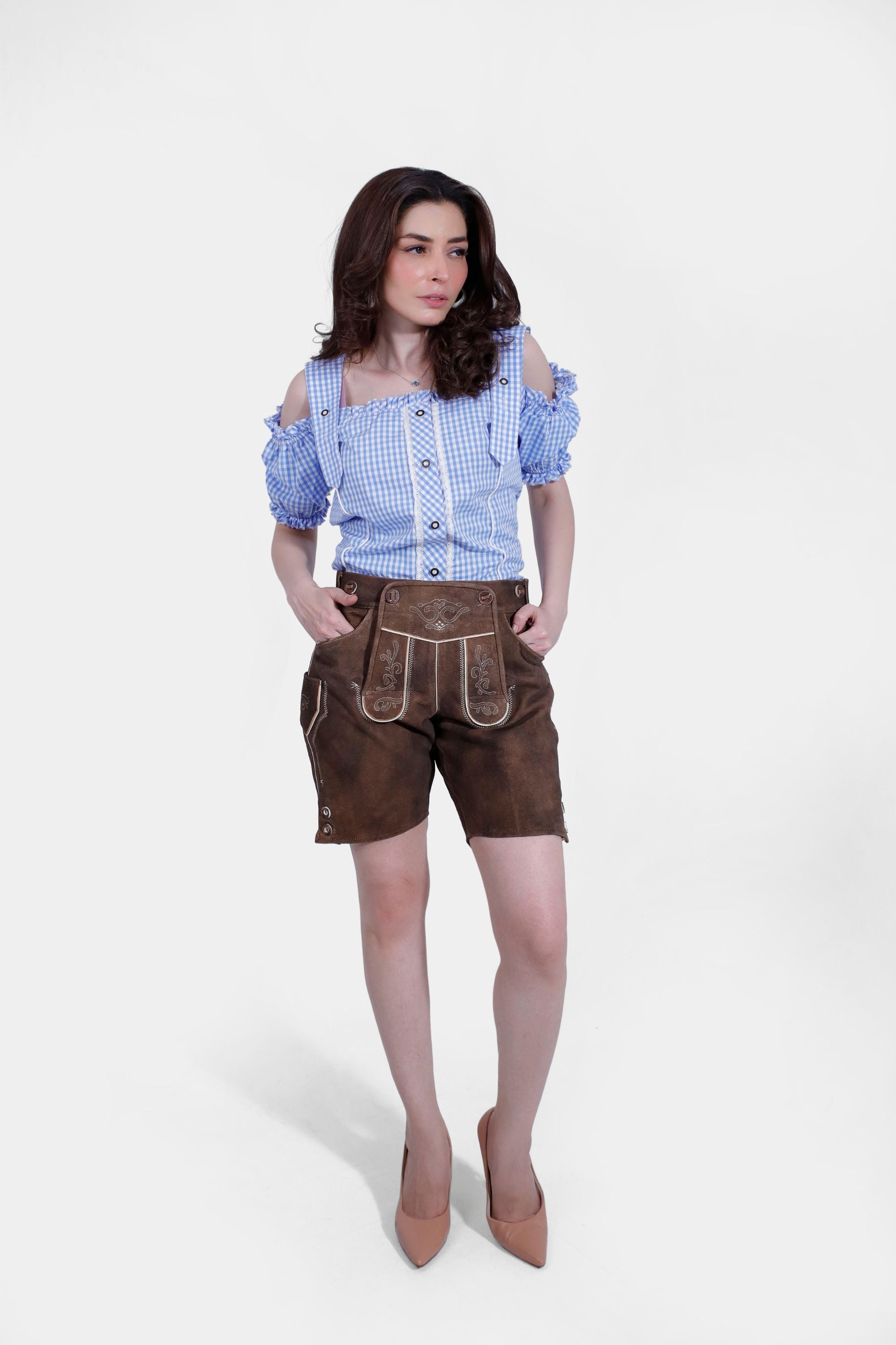 A woman wearing a blue and white checkered off-shoulder blouse and brown short lederhosen. She stands with hands in pockets, showcasing the traditional Bavarian outfit.