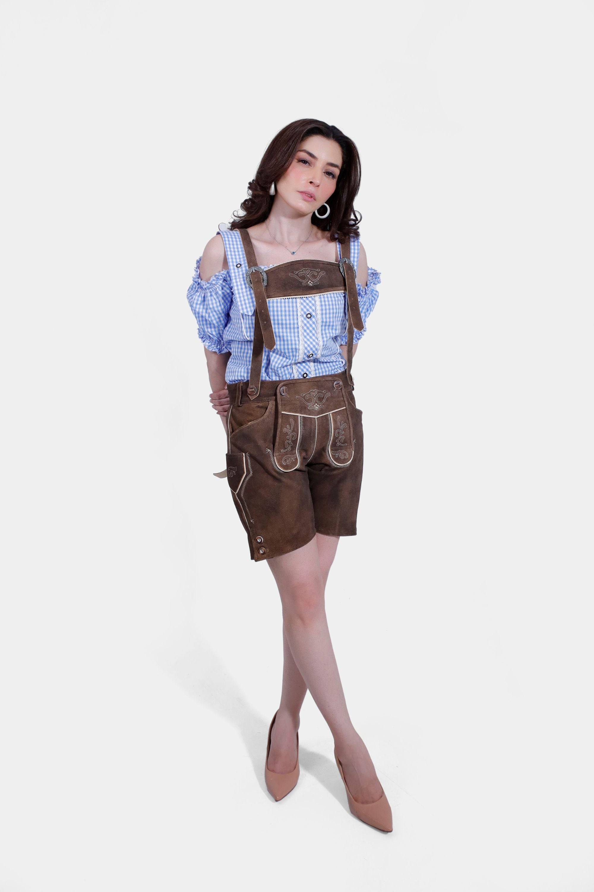 A woman wearing a blue and white checkered off-shoulder blouse and brown short lederhosen with suspenders. She stands with hands behind her back, showcasing the traditional Bavarian outfit in full view.