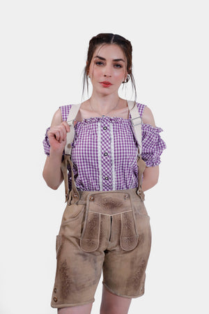 A close-up view of a woman in a purple checkered off-shoulder blouse and tan leather lederhosen shorts. She is holding the straps of her lederhosen and looking at the camera with a neutral expression