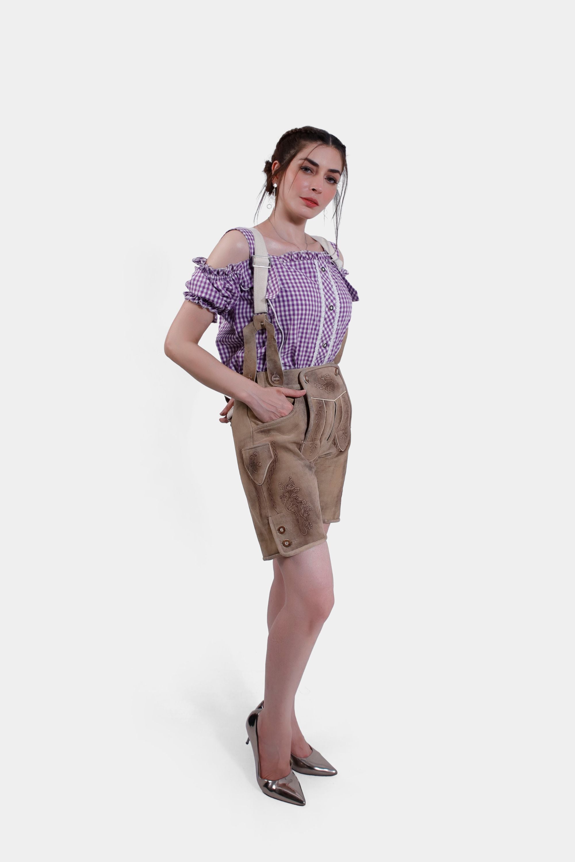 full-body side view of a woman wearing a purple checkered off-shoulder blouse and tan leather lederhosen shorts. She is posing with her hand in one pocket, and she is wearing metallic high heels.