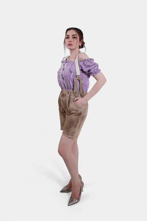 A full-body image of a woman modeling beige Lederhosen with suspenders and a purple checkered blouse, showcasing the traditional Bavarian outfit.