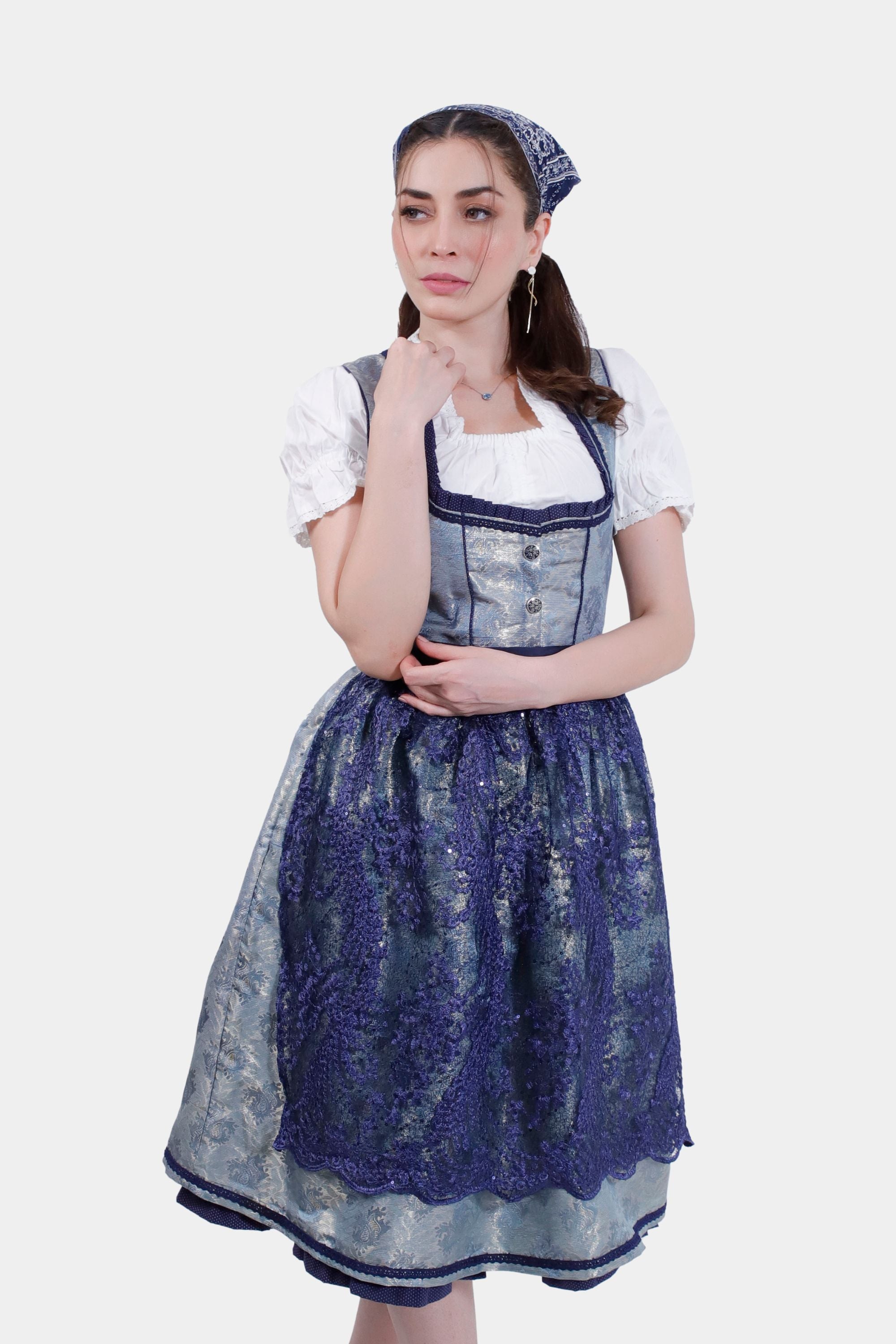 Woman wearing a traditional Dirndl dress in silver and blue, with a matching headscarf, posing with hand resting under her chin.