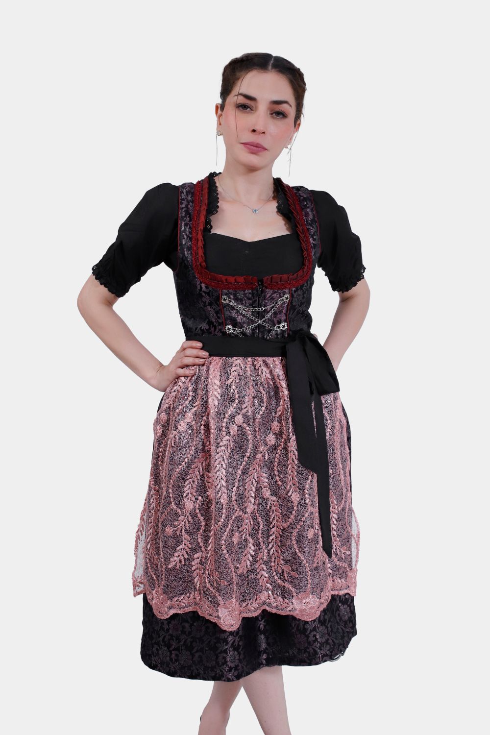 A woman models a traditional dirndl dress with a black bodice, red trim, and a lace pink apron over a floral-patterned black skirt.