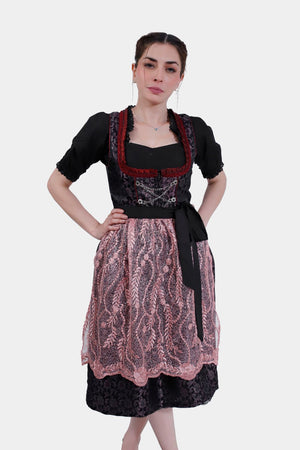 A woman models a traditional dirndl dress with a black bodice, red trim, and a lace pink apron over a floral-patterned black skirt.