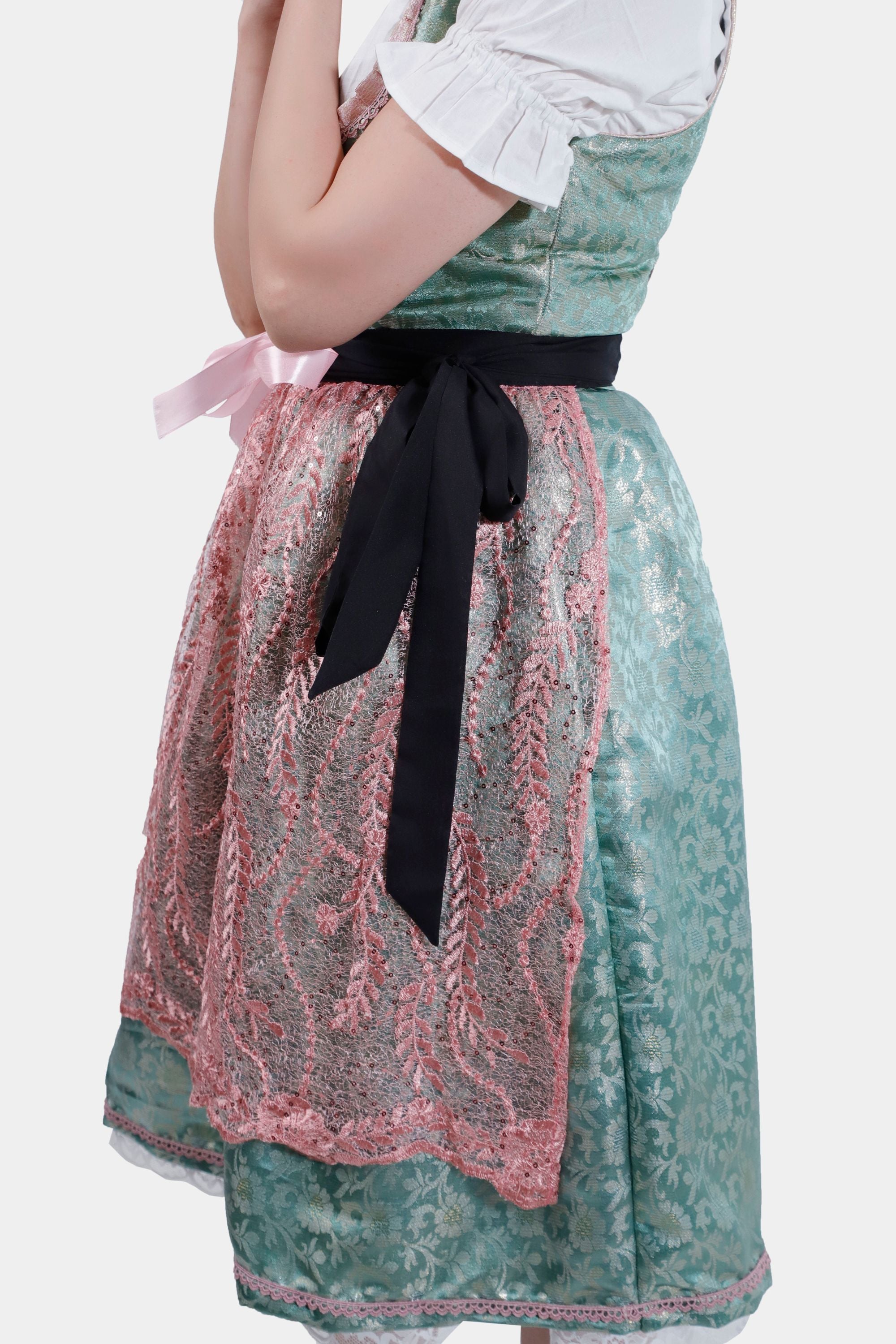 A close-up side view of the dirndl, highlighting the intricate pink lace apron and the black ribbon tied at the waist. The woman’s arm is bent, with her hand near her face.