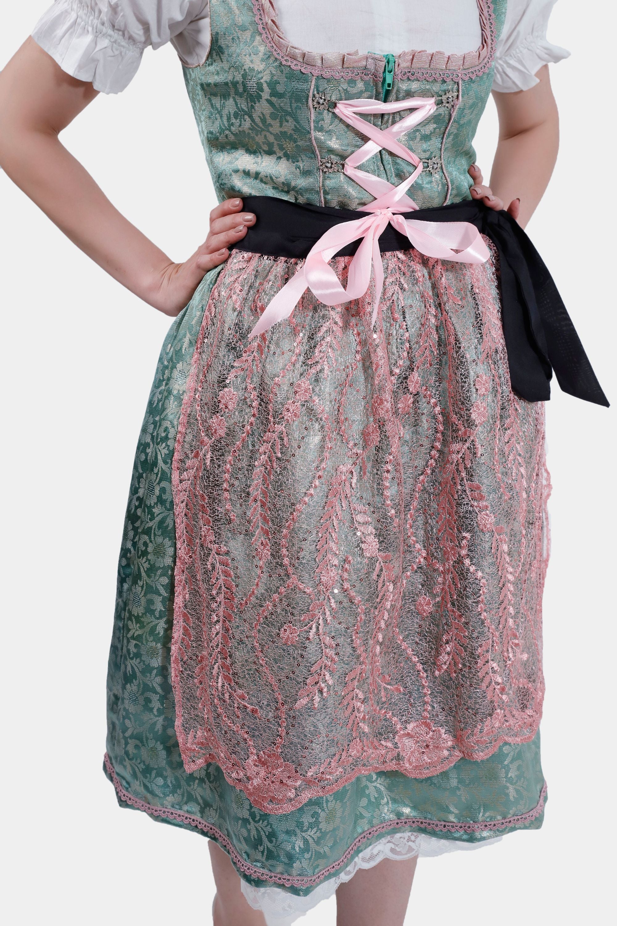 A detailed shot of the dirndl, focusing on the pink lace apron and the green bodice with pink lace-up detail. The black ribbon at the waist adds contrast to the outfit.