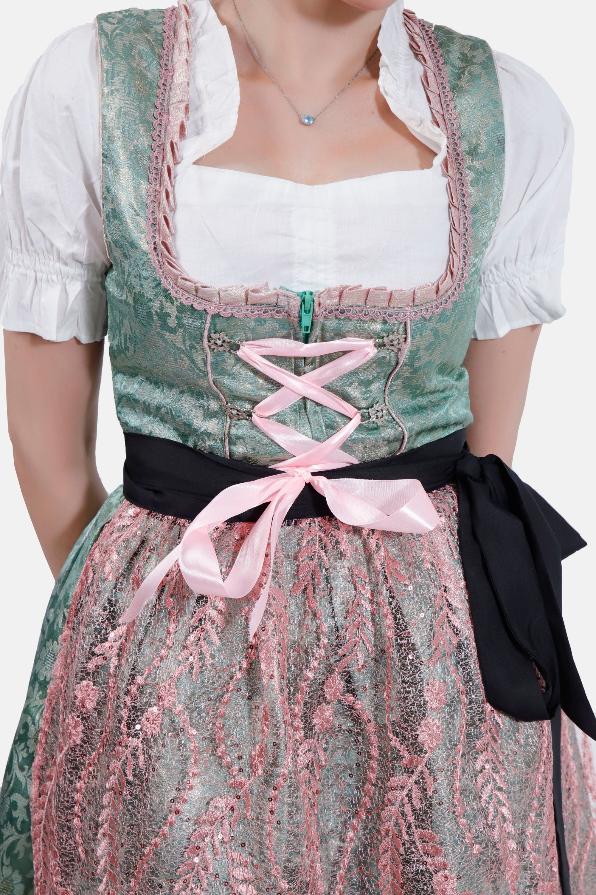 A close-up view of the dirndl, highlighting the intricate pink lace apron and the black ribbon tied at the waist. The woman’s arm is bent, with her hand near her face.