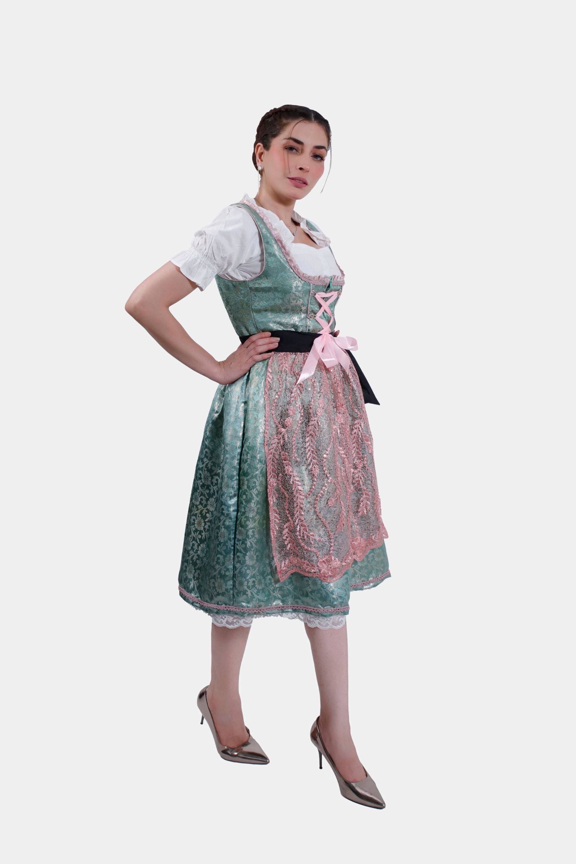 A full-length side profile of a woman wearing a green dirndl with pink lace accents, standing in metallic pointed heels. She has one hand on her hip and is looking off to the side, showcasing the detailed design of the dirndl's apron and the overall elegant look of the outfit.