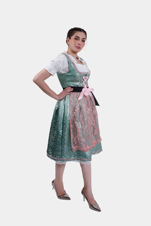A full-length side profile of a woman wearing a green dirndl with pink lace accents, standing in metallic pointed heels. She has one hand on her hip and is looking off to the side, showcasing the detailed design of the dirndl's apron and the overall elegant look of the outfit.