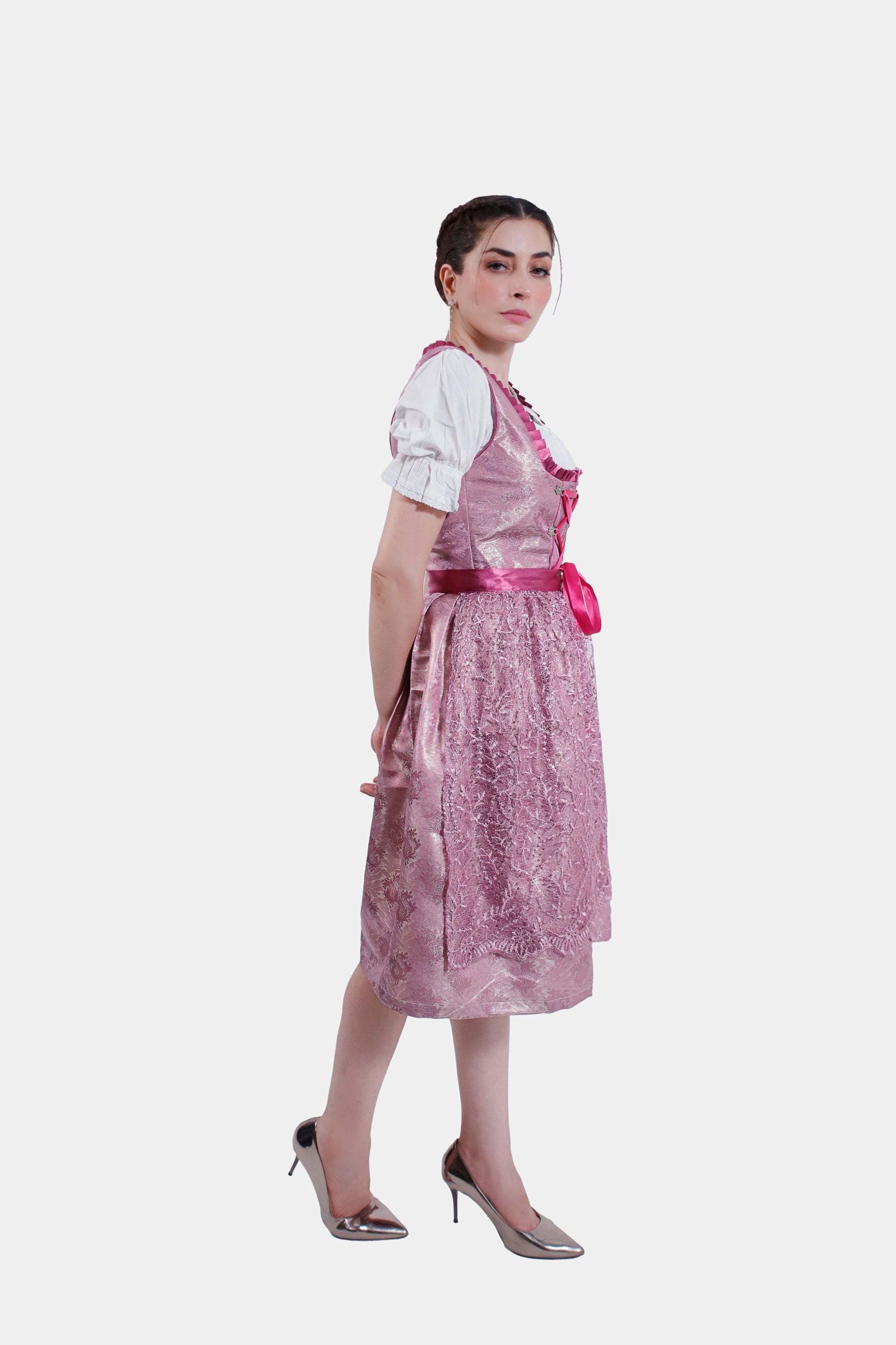 Side profile of the model in the pink dirndl, with a focus on the flow of the skirt and the lace and satin detailing, paired with metallic heels