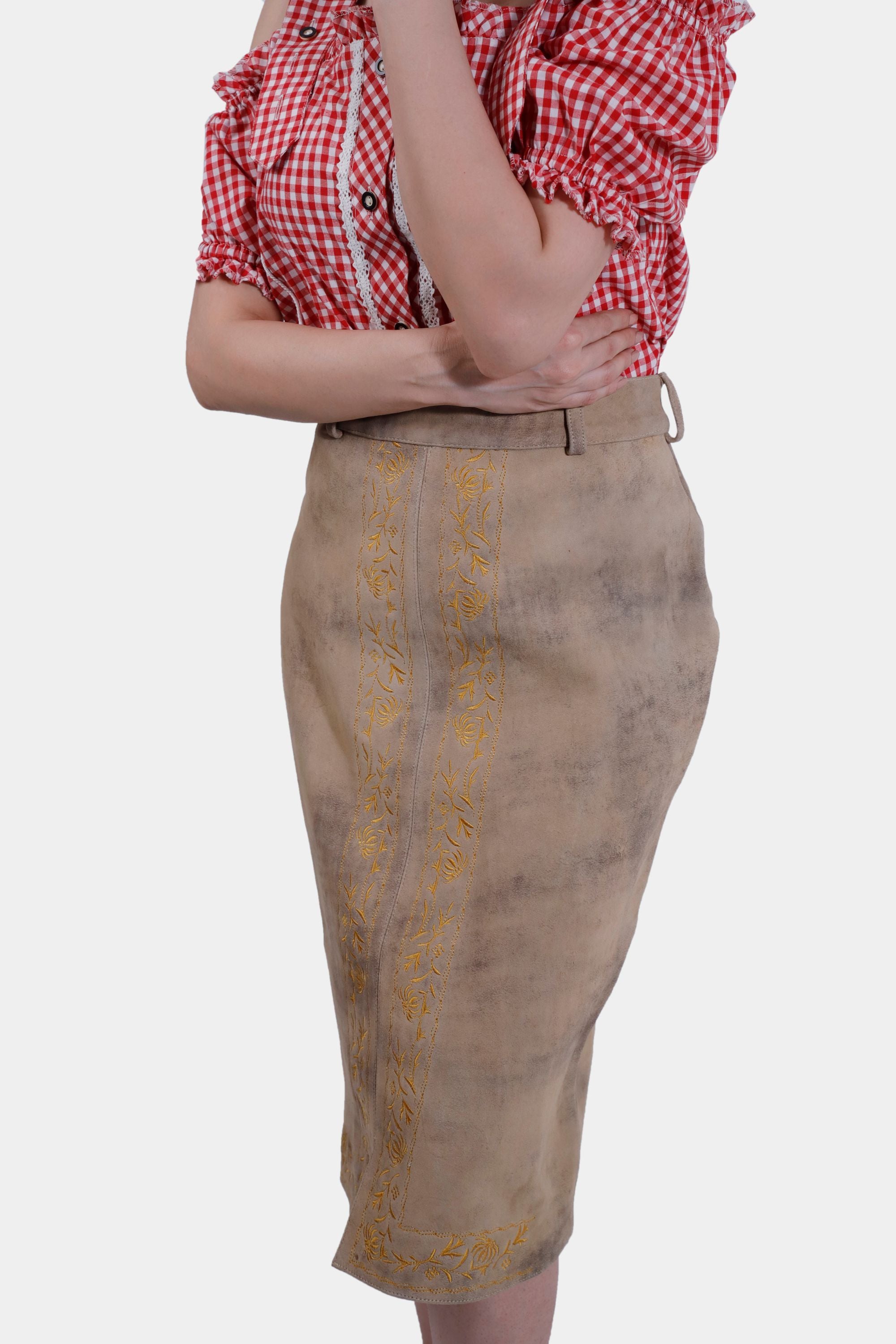 Side view close-up of the beige leather skirt with yellow embroidery. The woman's red and white checkered blouse with ruffled sleeves is partially visible as she crosses her arms.