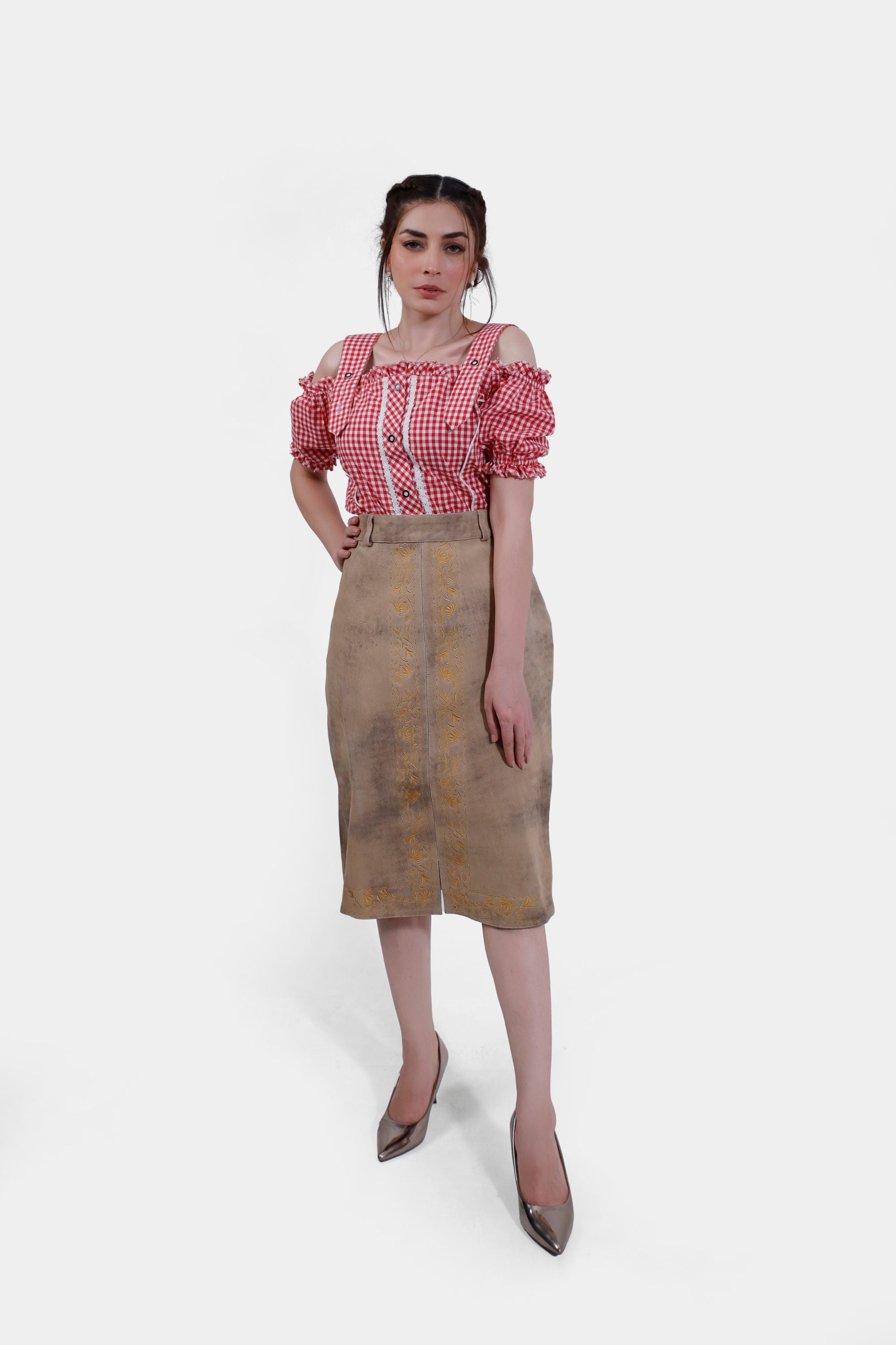 Full body of a woman in a red and white checkered blouse with off-shoulder ruffled sleeves and a beige leather skirt adorned with yellow embroidery. She is wearing metallic high heels and standing confidently.