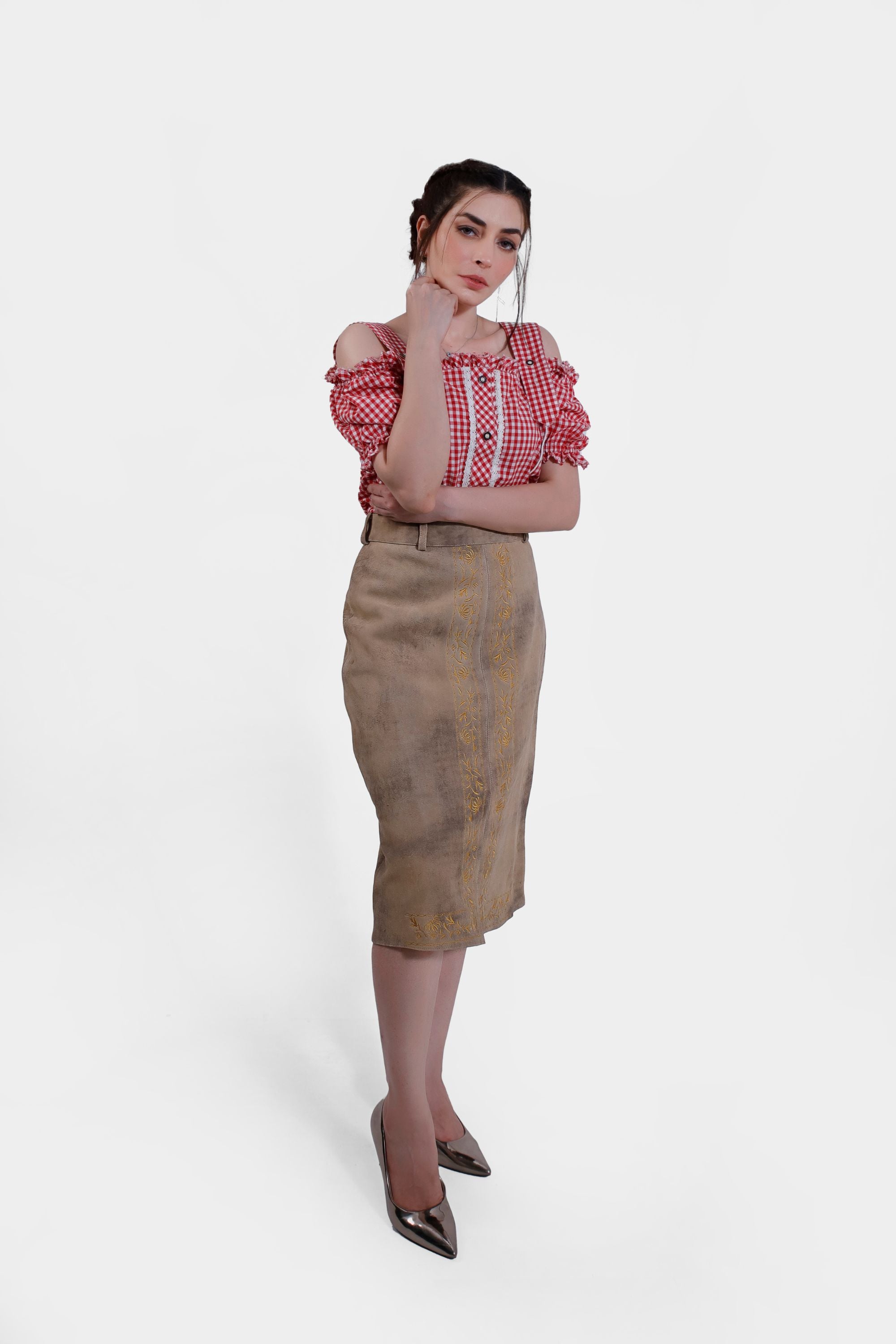 Full body shot of a woman wearing a red and white checkered blouse and a beige leather skirt with yellow embroidery. She stands with one hand under her chin, exuding a thoughtful pose.