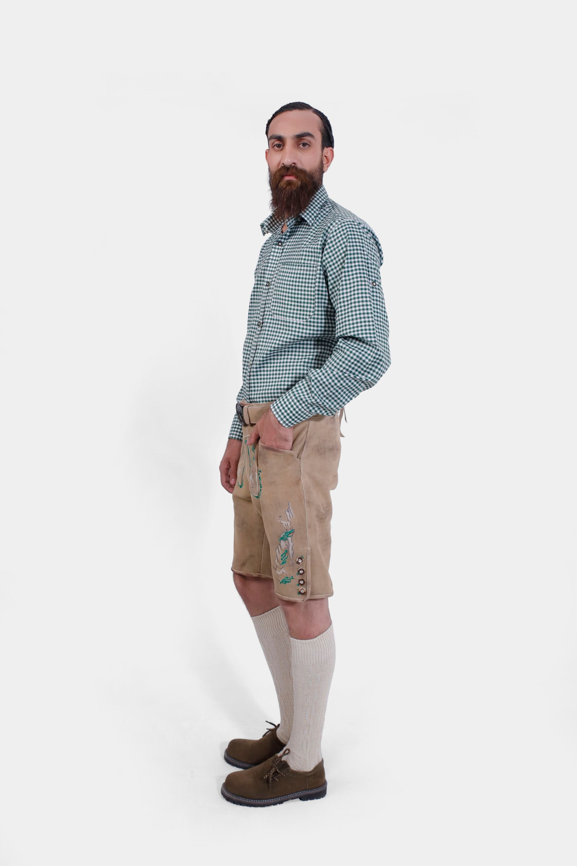 Side profile of a man with a beard wearing a green checkered shirt and beige lederhosen, hands in pockets, showing the side embroidery.