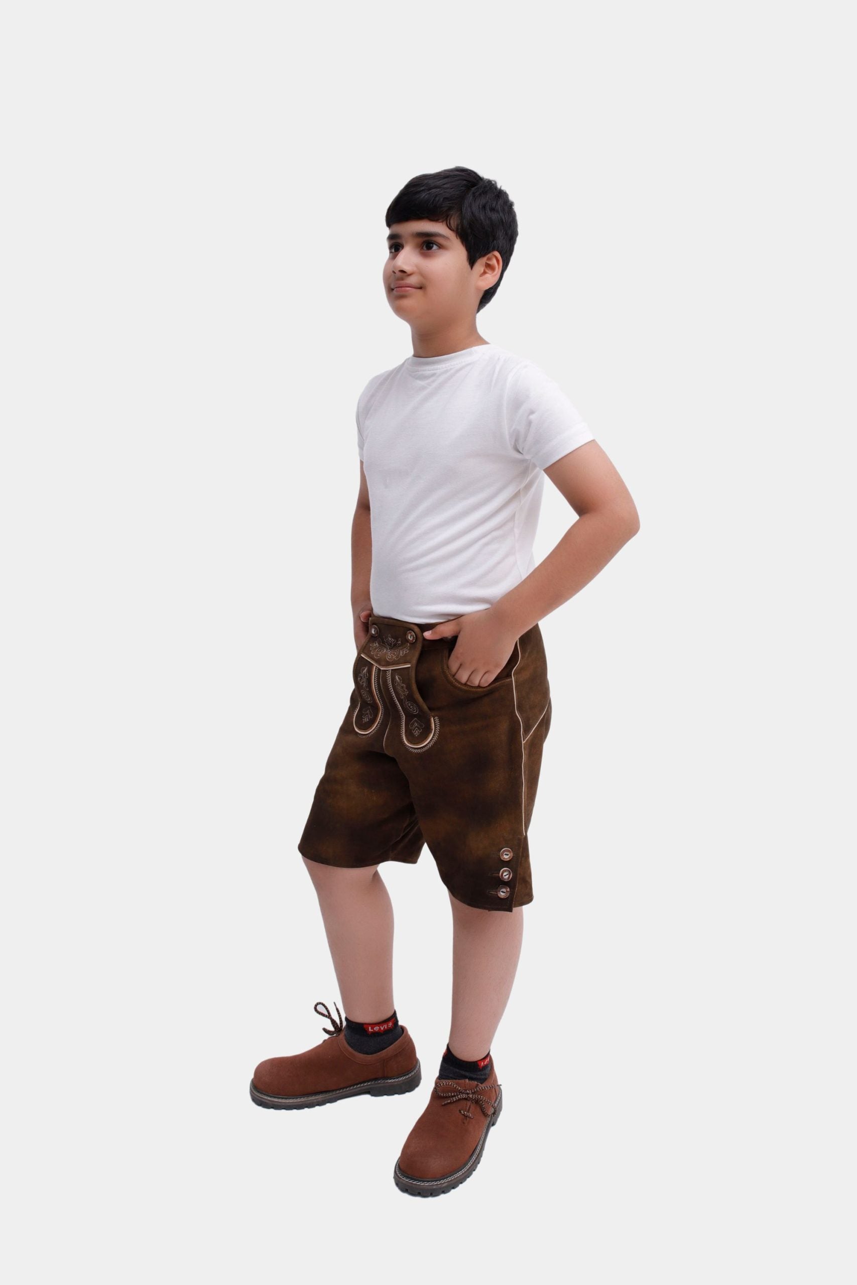 A young boy wearing a white T-shirt, brown embroidered Lederhosen, and brown shoes with black socks, standing sideways with his hands in his pockets against a white background.