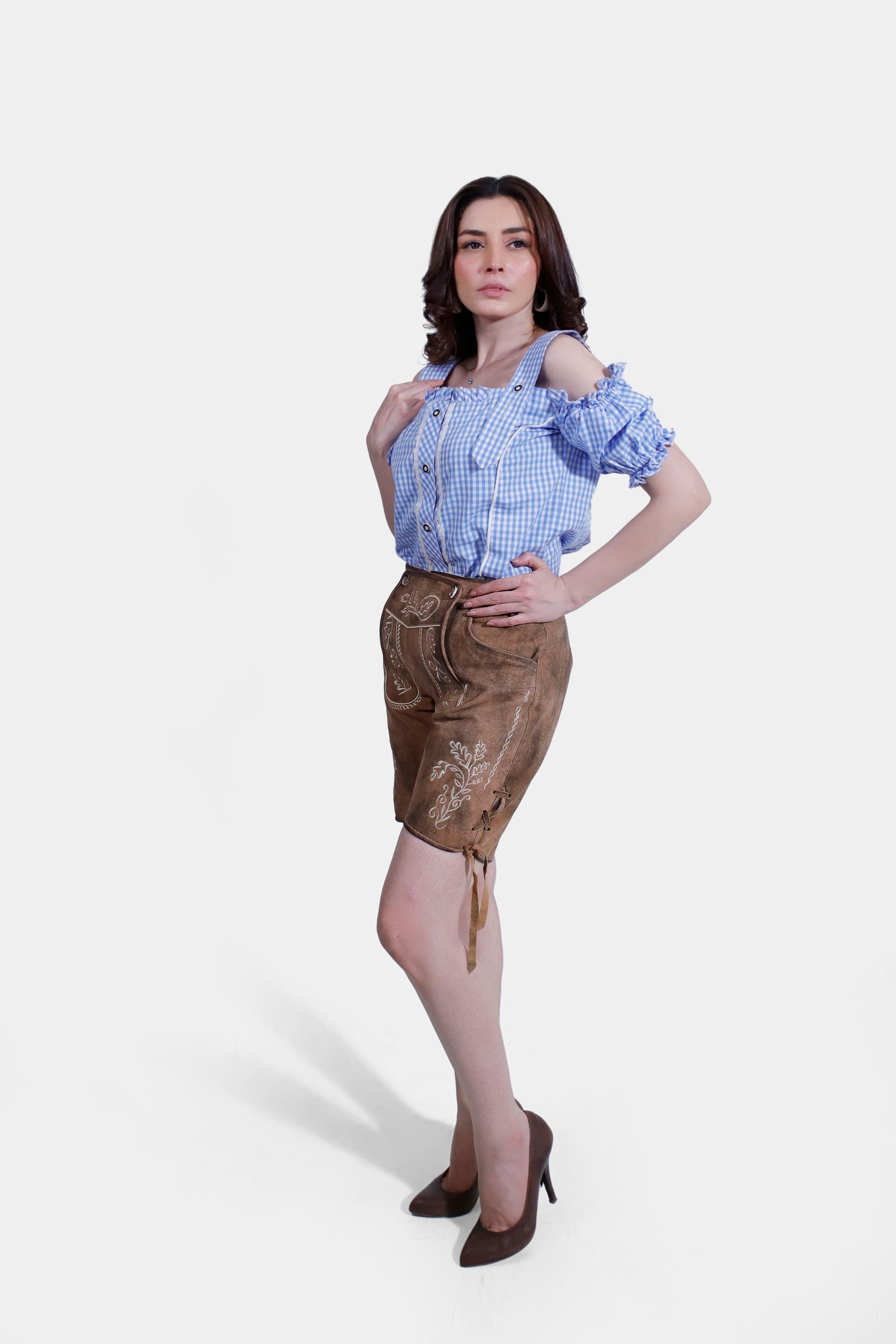 A woman in a blue checkered blouse and embroidered brown leather shorts, viewed from the side, posing with one hand near her chest.







