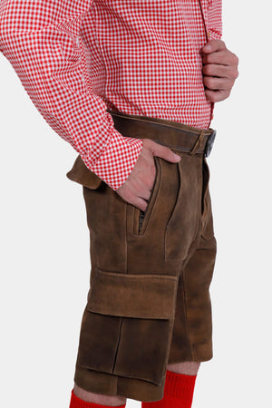 A full side view of the man wearing the brown leather lederhosen, standing with his hands in his pockets and facing to the side.