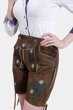 A side view of a woman in earthy brown lederhosen with green and white floral embroidery, paired with a white shirt, posing with her hand on her hip.