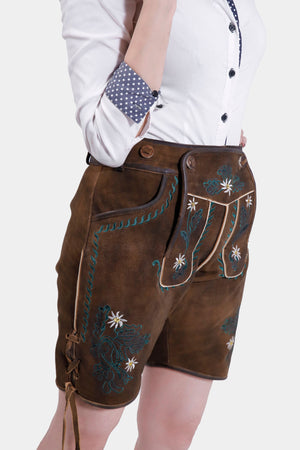 Close-up of a model wearing brown suede traditional Bavarian leather shorts with intricate green and white floral embroidery, paired with a white button-up shirt featuring polka-dotted cuffs.