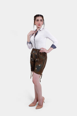 Full body view of a model wearing brown suede traditional Bavarian leather shorts with green and white floral embroidery, styled with a white button-up shirt and nude high heels.