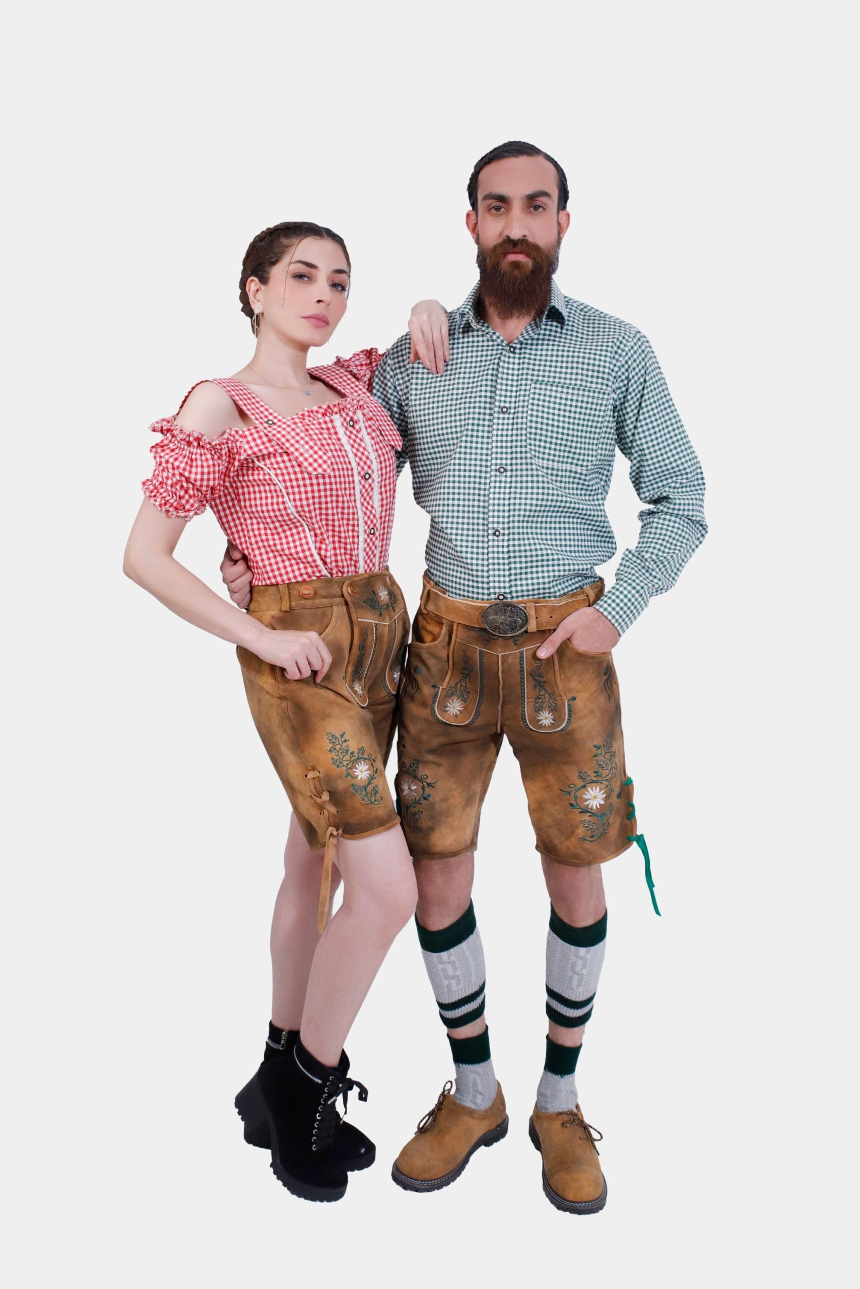A couple posing in matching Freude Couple Lederhosen outfits. The man wears a green checkered shirt and Lederhosen shorts, while the woman sports a red checkered off-shoulder blouse and Lederhosen shorts