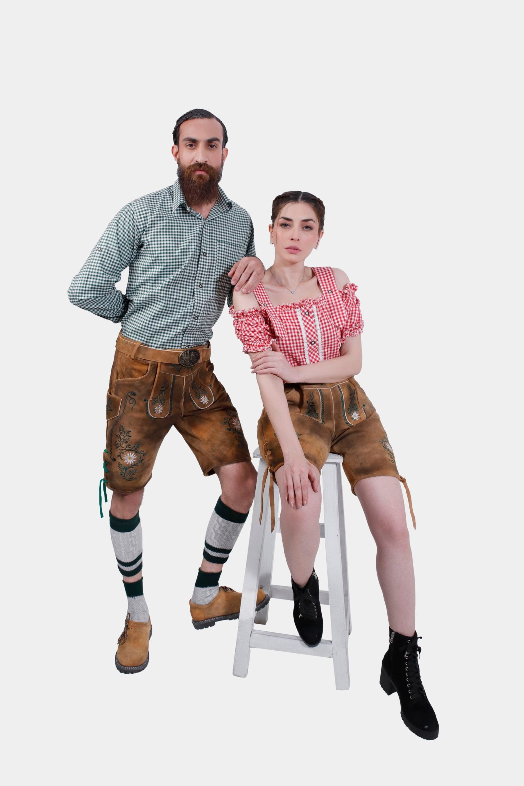 A couple in Freude Couple Lederhosen outfits, with the man leaning on a stool in a green checkered shirt and Lederhosen shorts, and the woman sitting on the stool in a red checkered off-shoulder blouse and Lederhosen shorts.