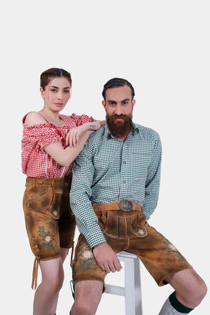 A couple wearing matching Freude Couple Lederhosen outfits. The man is seated wearing a green checkered shirt and Lederhosen shorts, while the woman stands beside him in a red checkered off-shoulder blouse and Lederhosen shorts.