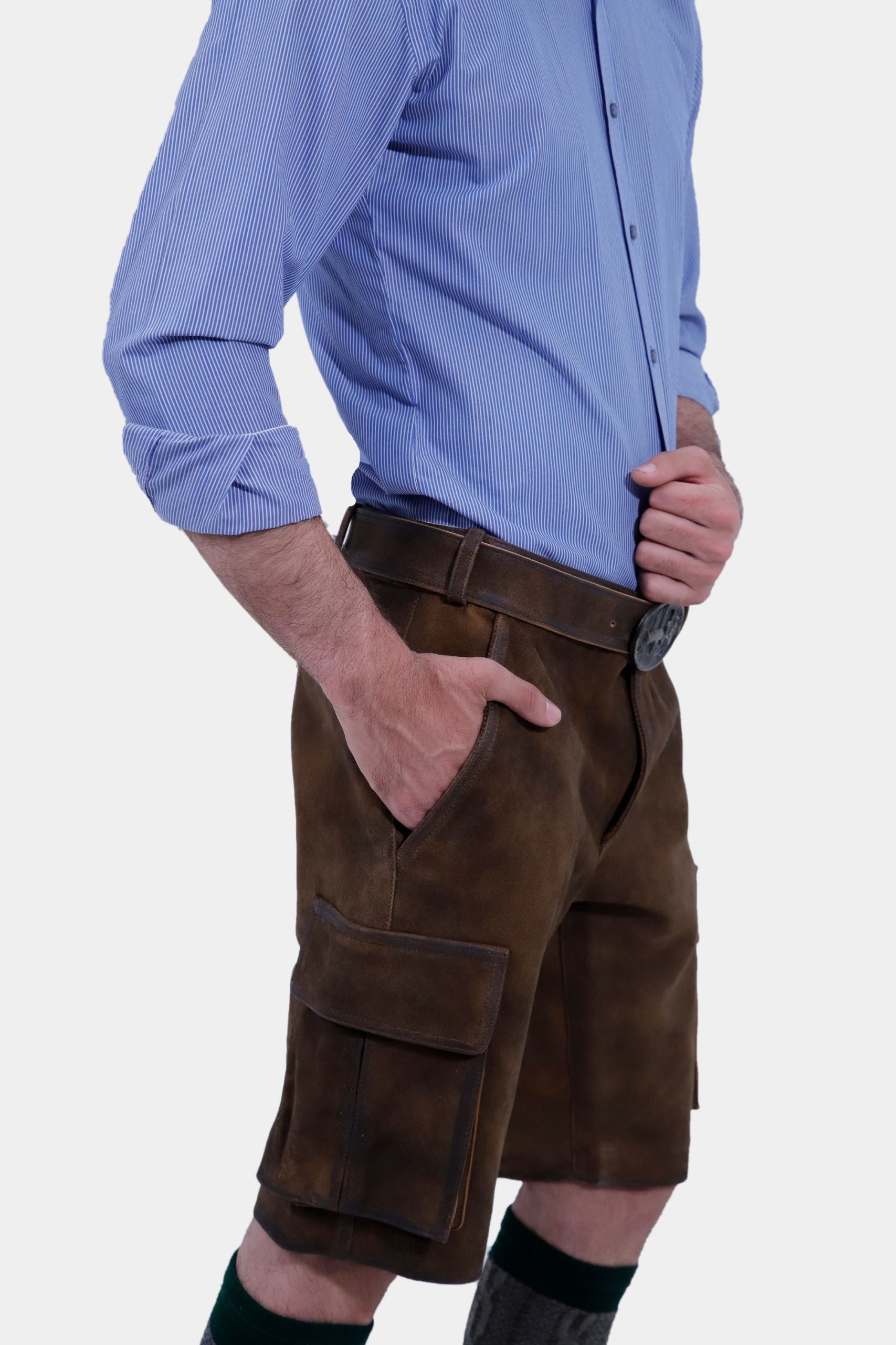 Closed Side view of a man in a blue striped shirt is posing in traditional brown leather cargo Lederhosen. The outfit is accessorized with a belt and traditional Bavarian socks and shoes. The side view showcases the side pockets and the decorative belt buckle.
