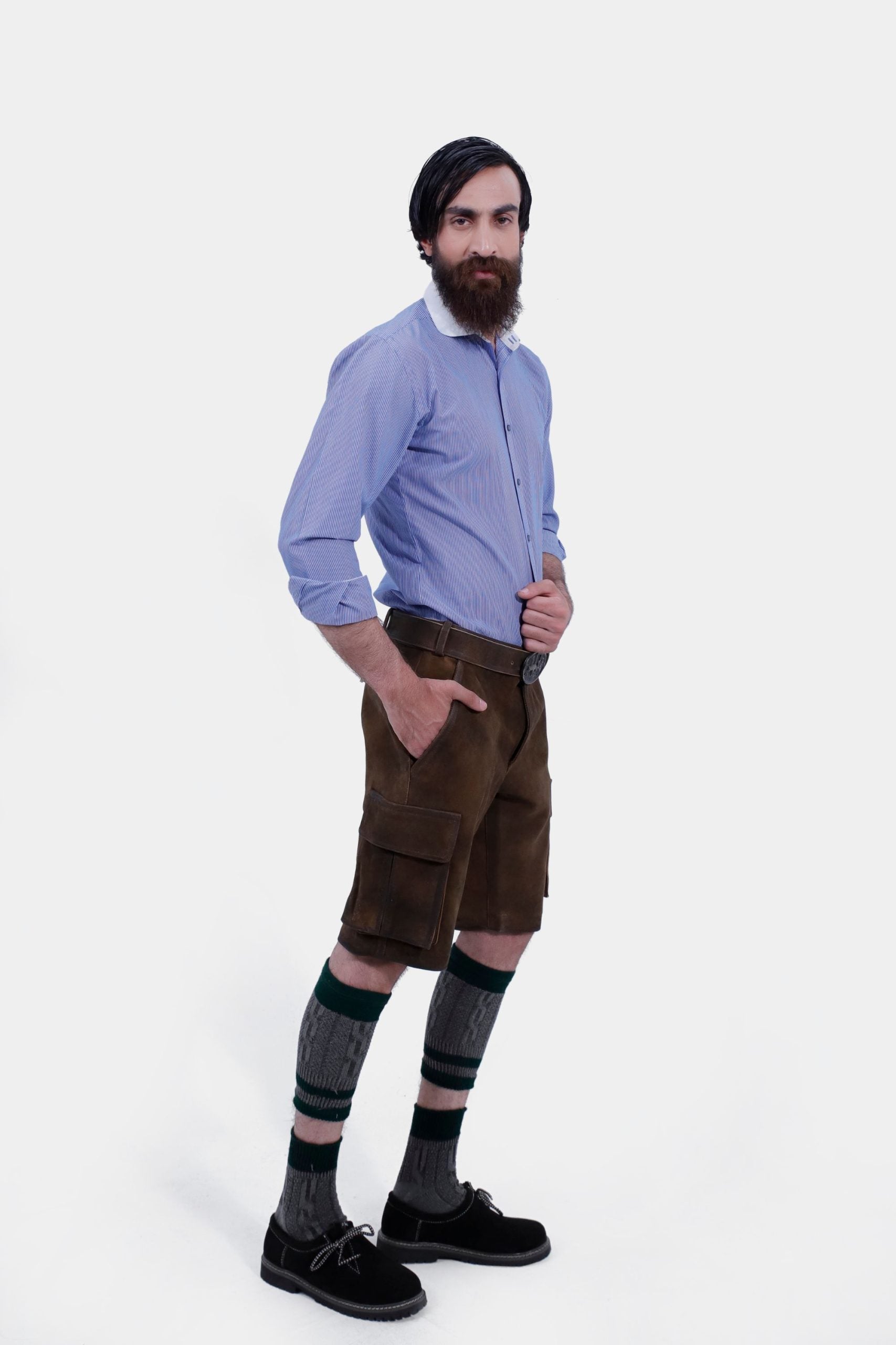 A man in a blue striped shirt is posing in traditional brown leather cargo Lederhosen. The outfit is accessorized with a belt and traditional Bavarian socks and shoes. The side view showcases the side pockets and the decorative belt buckle.