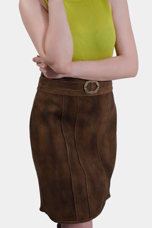A close-up side view of the brown leather skirt, highlighting its detailed stitching and the ornate circular buckle on the belt. The woman's arm is partially visible, crossed over her body, emphasizing the texture and fit of the skirt.