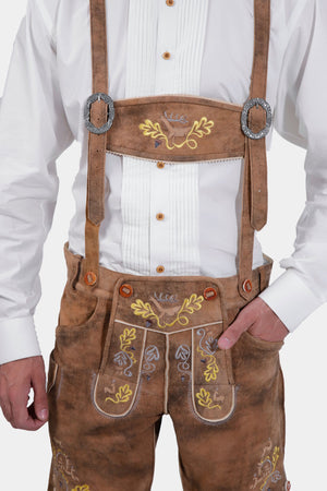 A close-up view of a man wearing traditional lederhosen with Gerd Muller suspenders. The suspenders feature intricate embroidery and detailed metal buckles, complementing the rustic brown suede lederhosen adorned with yellow and purple floral embroidery.