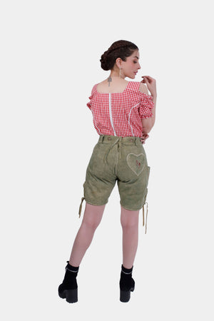 A woman with braided hair wearing a red and white checkered off-the-shoulder top and green lederhosen with a heart design on the back pocket. She stands with her back to the camera, looking over her right shoulder.
