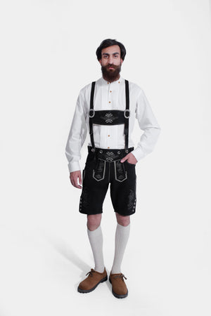 A man wearing a traditional Hardy Kruger Lederhosen outfit, featuring black Lederhosen shorts with white embroidery and a crisp white shirt, standing with a neutral expression.